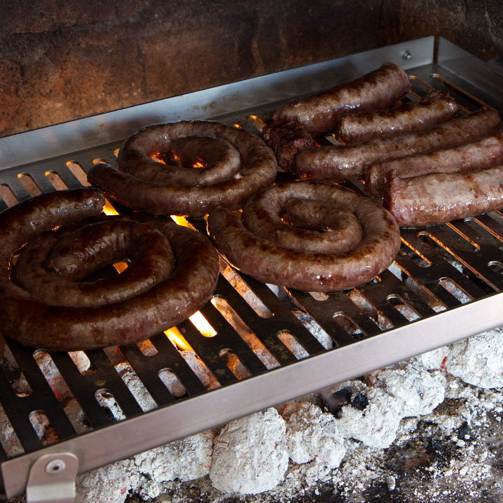 Sausages grilling on a stainless steel braai grid with flames and smoke.