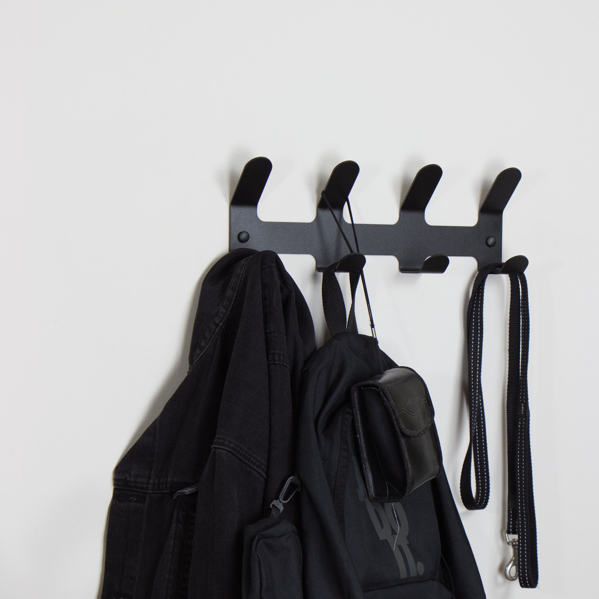 Black wall-mounted coat rack with hooks holding a jacket, bag, and leash on a white background